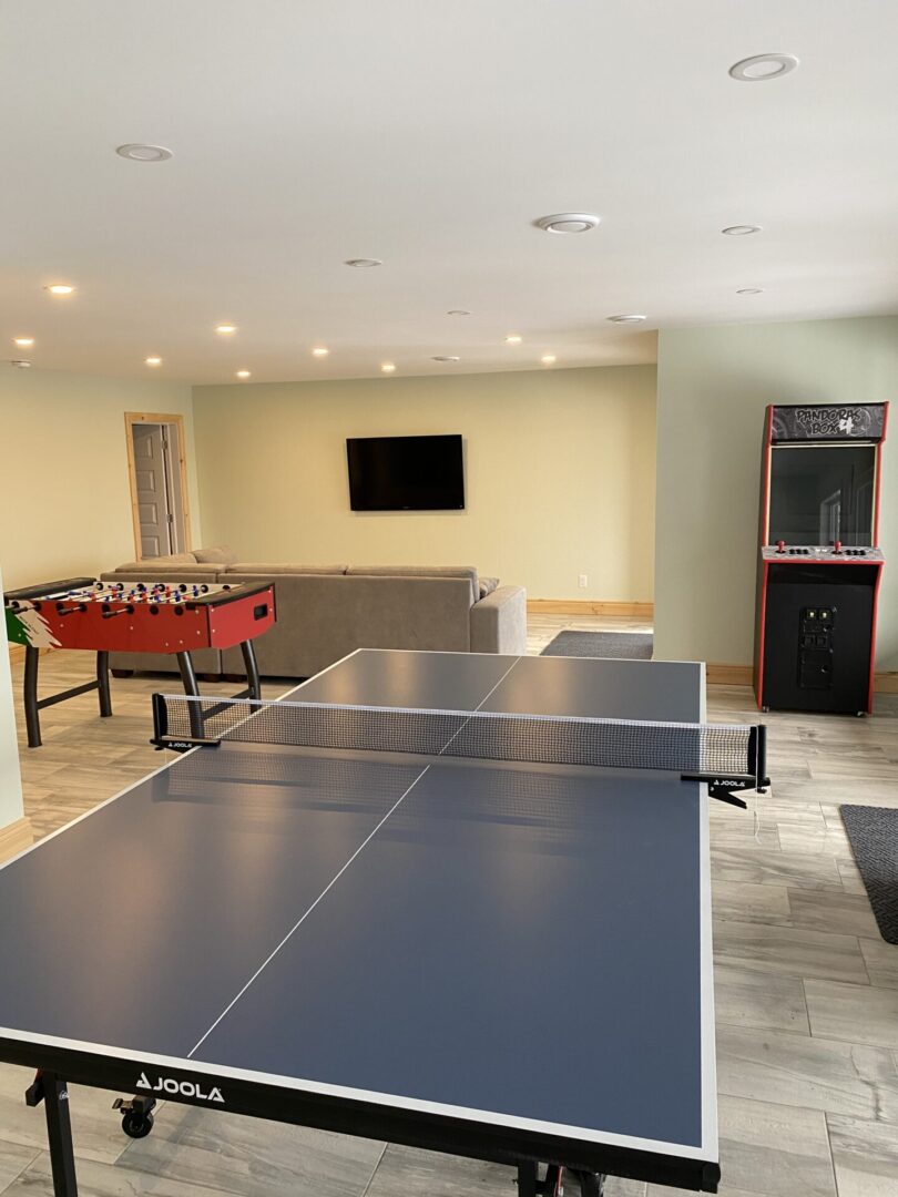 A game room with a ping pong table, foosball table, arcade machine, and TV.